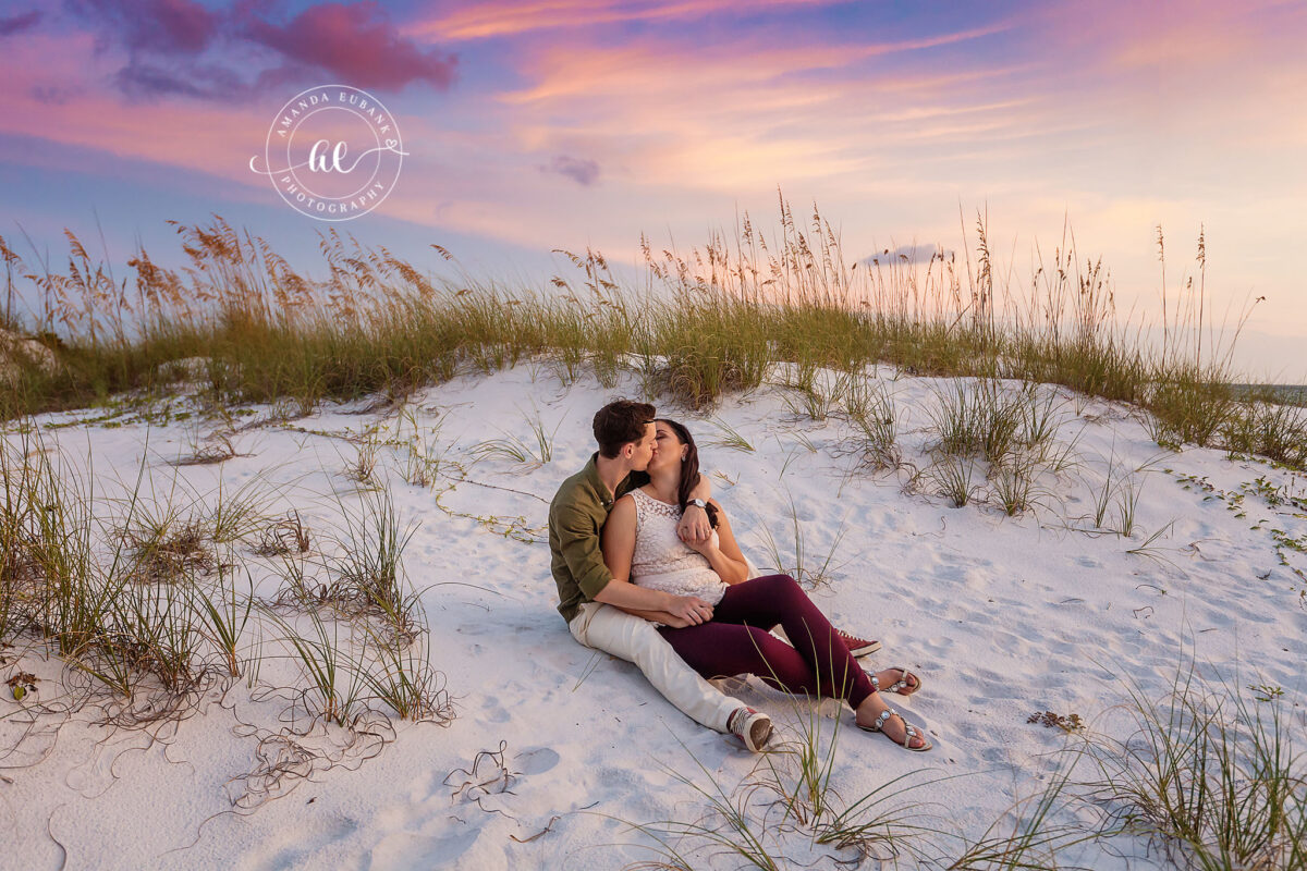 Destin engagement photographer captures couple kissing on sand dunes among sea oats at sunset