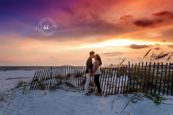 Destin engagement photographer captures couple kissing by beach fence at dramatic sunset