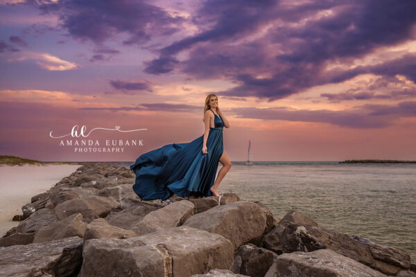 Destin senior photographer captures blonde in flowing navy gown on jetty boulders at purple sunset