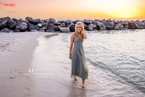 Destin senior photographer captures blonde senior in gray gown wading along jetty rocks at sunset