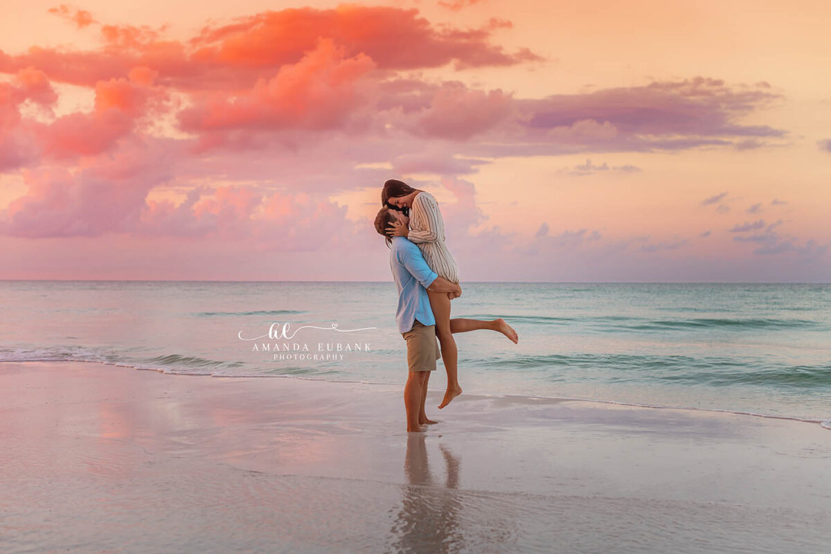 Destin engagement photographer captures couple lifted in joyful embrace at pink sunset on the beach